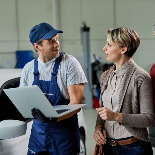 Smiling car mechanic using laptop while talking to a couple in repair shop.